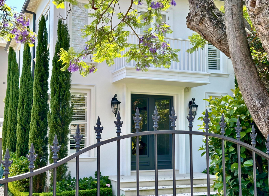 A white house with green door, tall trees, purple flowers, and a black wrought iron fence in front.