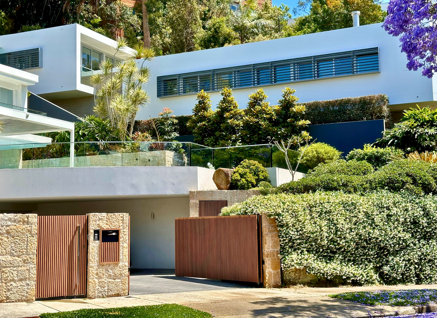 Modern white house with large windows, lush gardens, and a wooden gate on a sunny day.